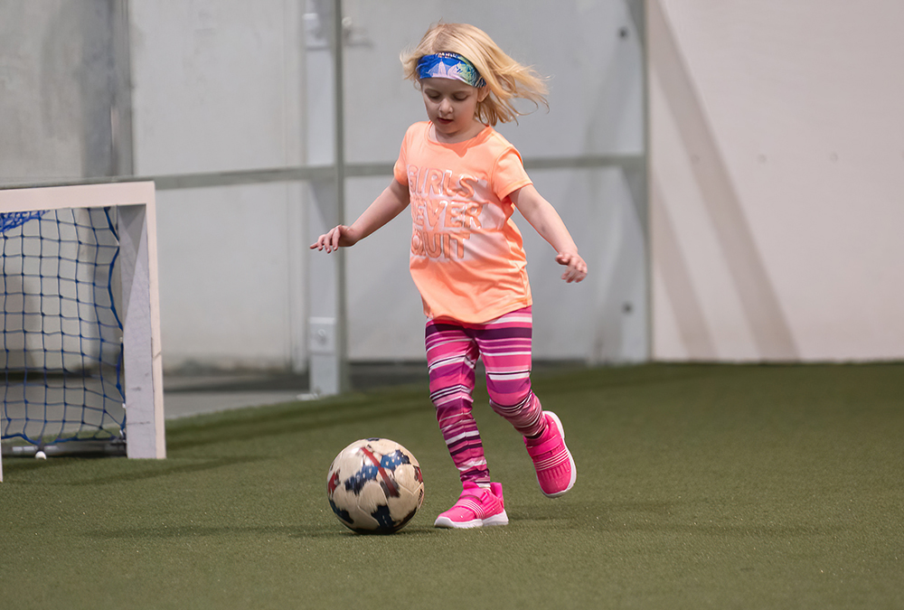 little girl in pink shirt and blue headband dribbling soccer ball