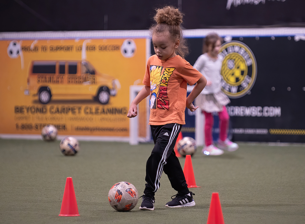 little girl in orange shirt dribbling soccer ball around cones