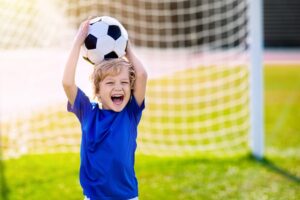 little boy in blue shirt with a huge smile balancing soccer ball on head