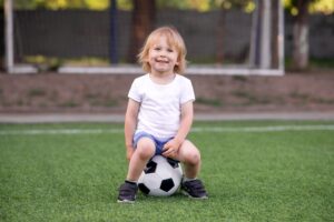 little boy sitting on soccer ball at edge of field