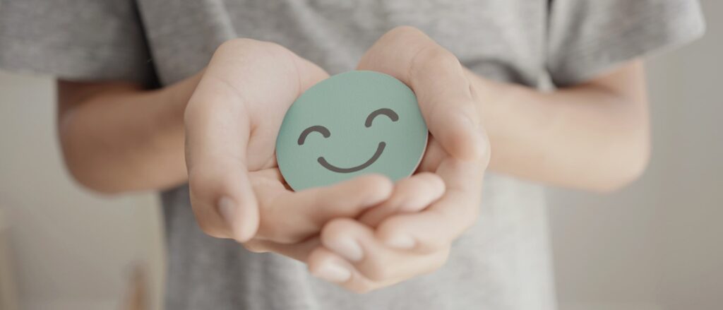 Teen holding a cardboard cutout of a smiley face, symbolizing positive mental health and emotional well-being.