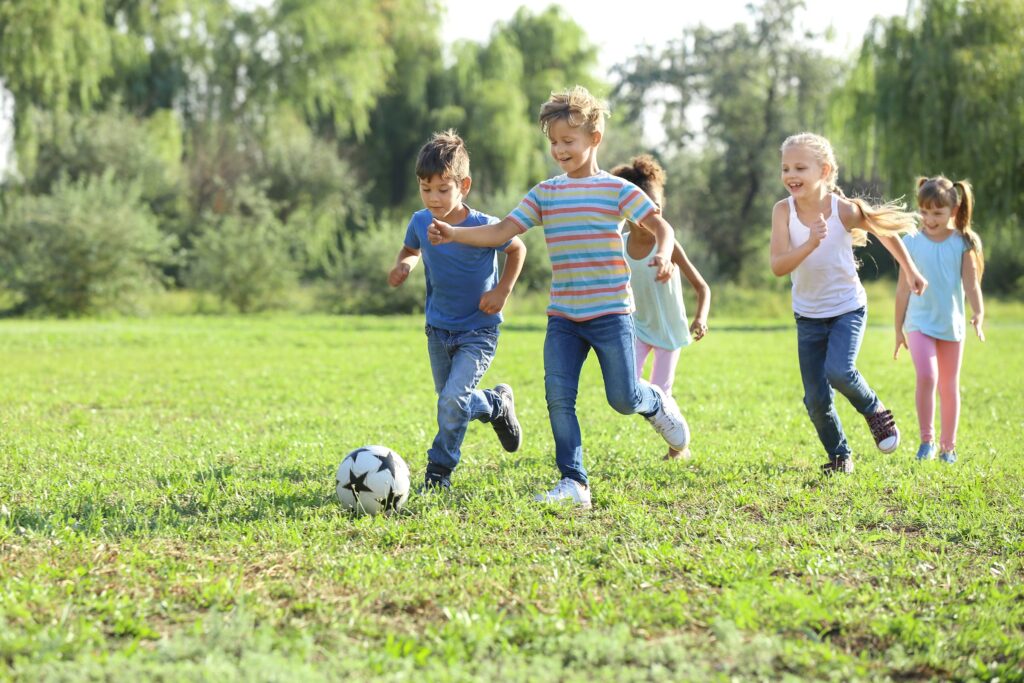 Children in casual street clothes chasing a soccer ball in a grassy park field during unstructured play.