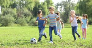 Children in casual street clothes chasing a soccer ball in a grassy park field during unstructured play.