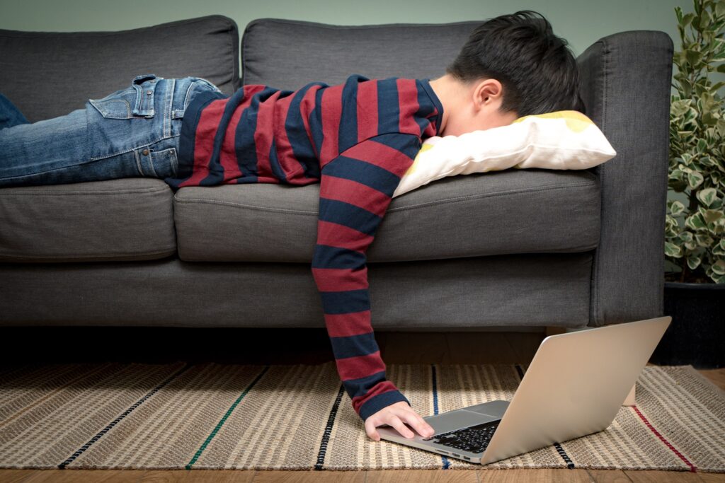 Child laying facedown on a couch, reaching toward a laptop on the floor, appearing overwhelmed by activities and responsibilities.