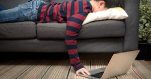 Child laying facedown on a couch, reaching toward a laptop on the floor, appearing overwhelmed by activities and responsibilities.