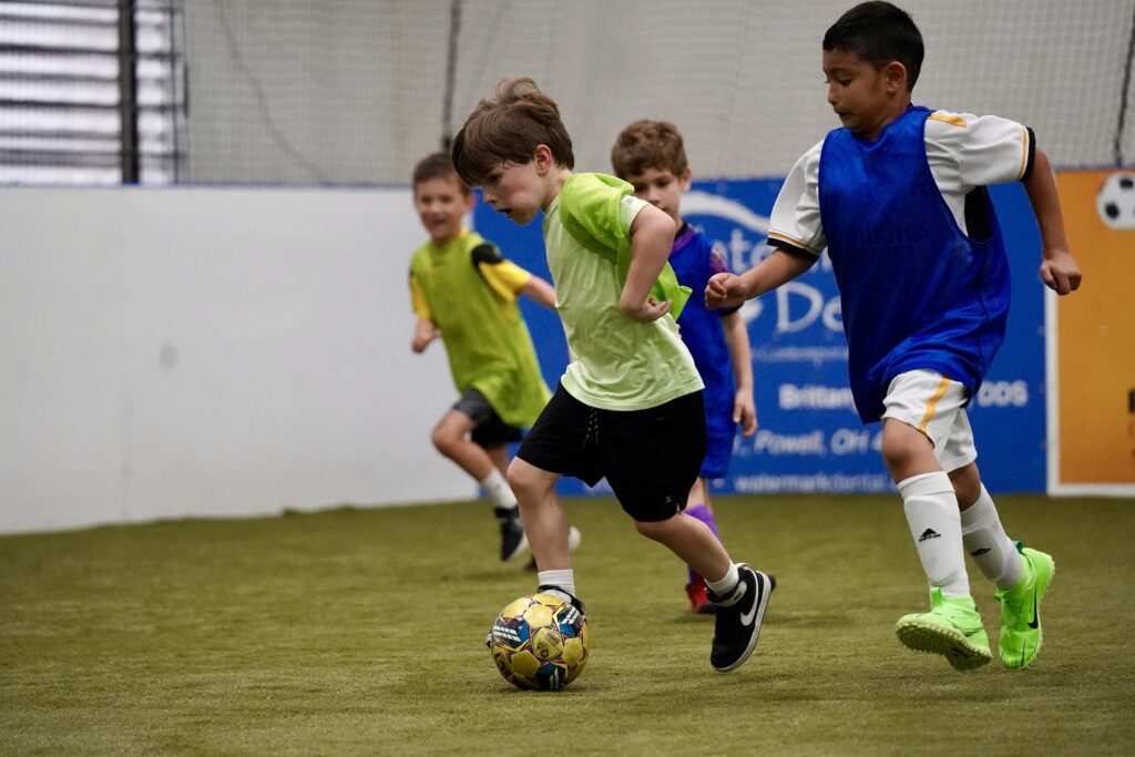 Youth soccer players scrimmaging in an indoor soccer facility during summer camp.