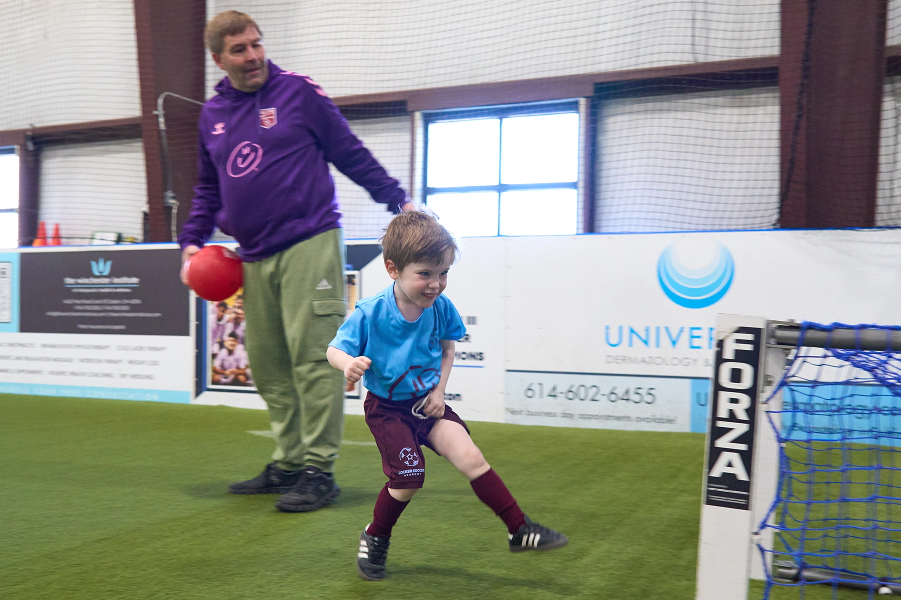 Coach holding a soccer ball behind a young boy who just kicked a ball into a small net during Little Kickers class at Locker Soccer Academy