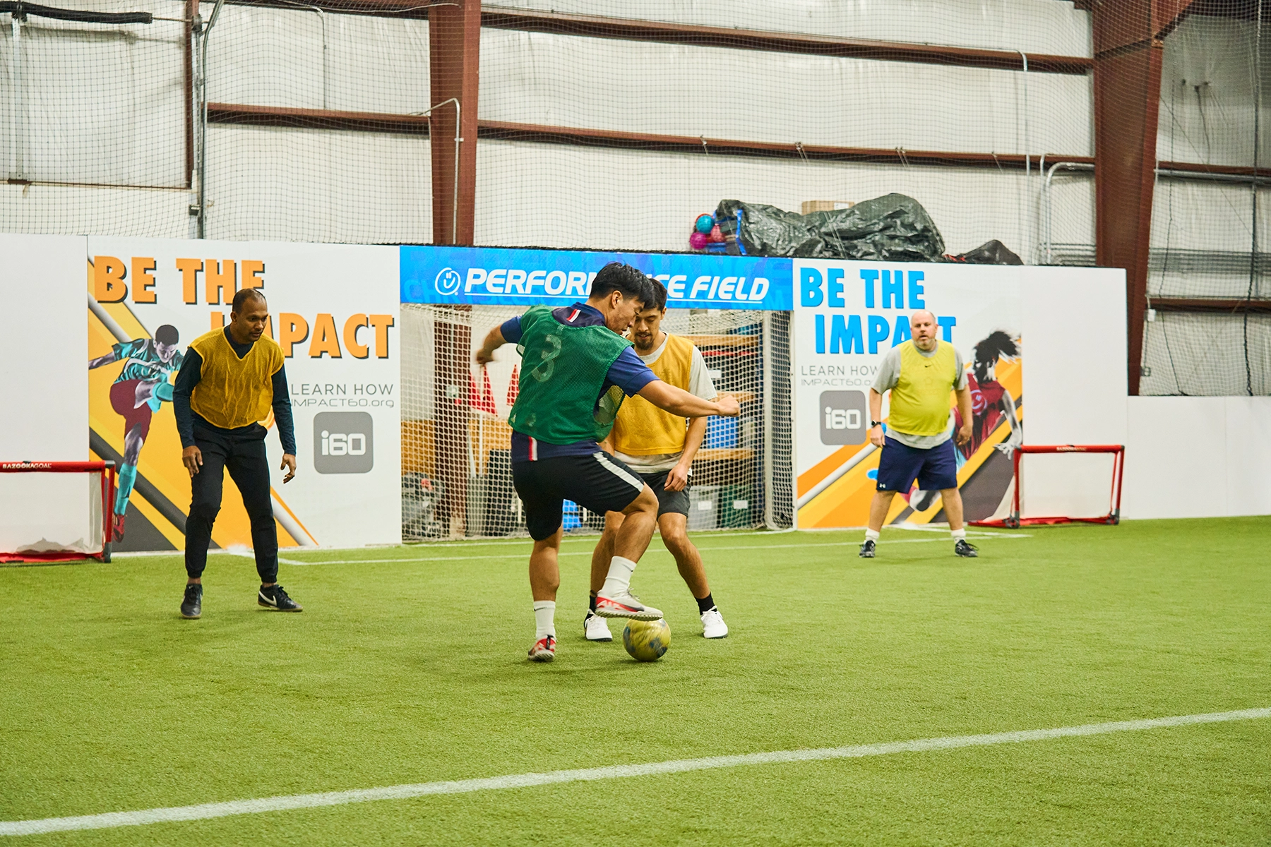 Adult pickup soccer game at Locker Soccer Academy with a green team player dribbling toward a yellow team defender indoors