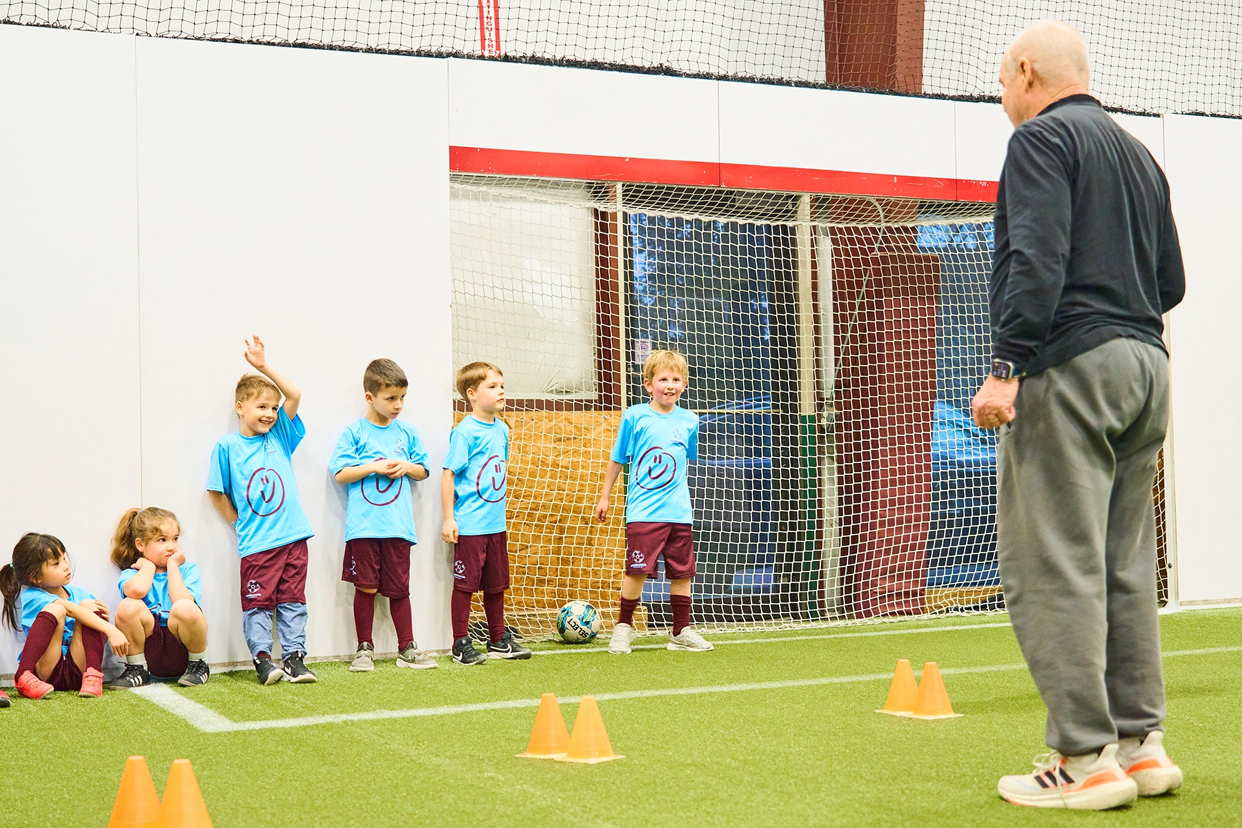 Coach Steve giving instruction to Elite Juniors players sitting and leaning on indoor field wall, one boy raising his hand to ask a question
