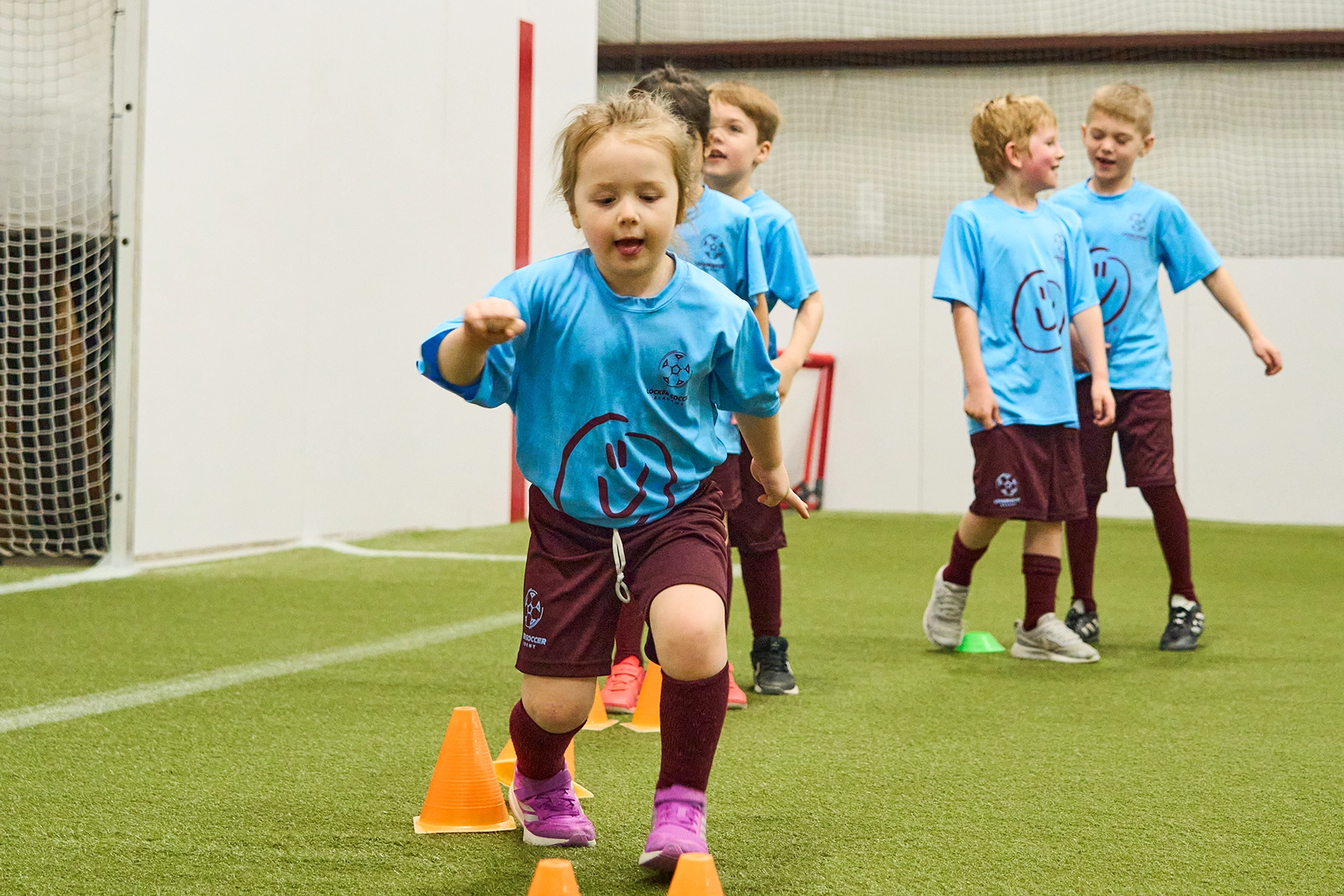 Kids in line performing a ball-free balance drill between cones at Locker Soccer Academy, with a young girl in motion at the front