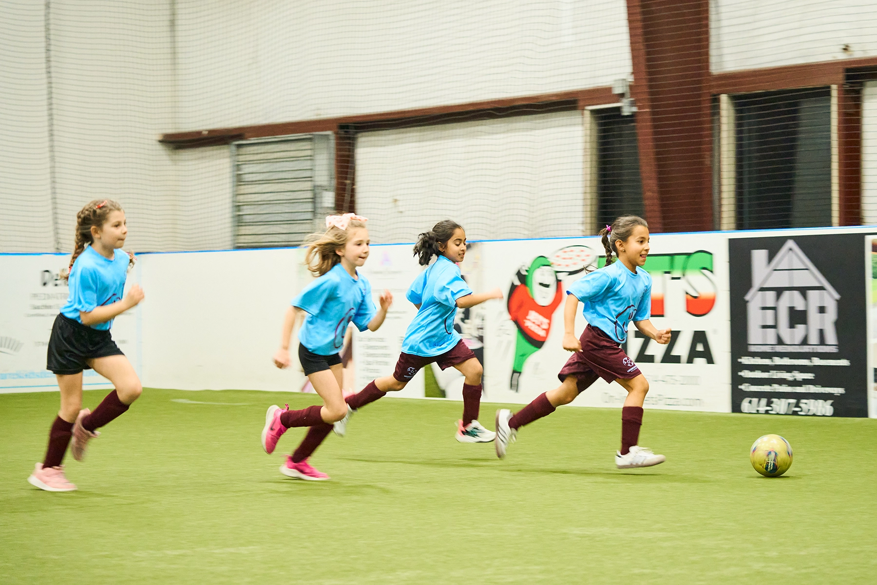 Four girls in uniform from Locker Soccer’s School of Excellence sprinting down the indoor field chasing the ball